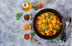 Top view of a bowl of chicken curry garnished with fresh parsley, surrounded by small bowls of spices, peppercorns, a garlic clove, a tomato, and a bottle of oil on a gray textured surface indian food recipes