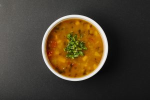 A bowl of lentil soup with vegetables and fresh herbs on top, served in a white bowl on a dark surface.
